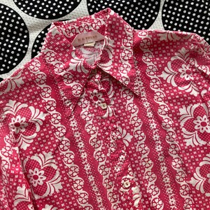 May include: A pink long-sleeved button-down shirt with a white floral pattern. The shirt has a collar and a button closure. The label on the shirt reads "M. & S. STANA."