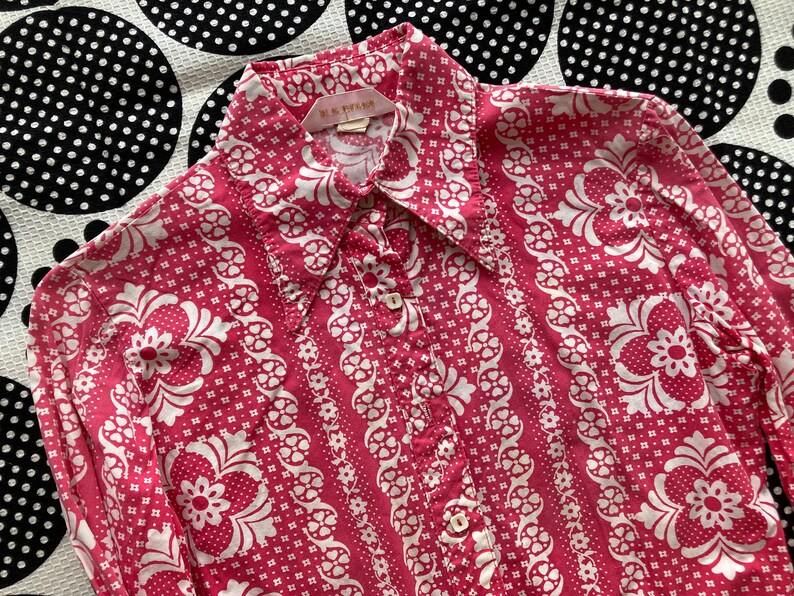 May include: A pink long-sleeved button-down shirt with a white floral pattern. The shirt has a collar and a button closure. The label on the shirt reads "M. & S. STANA."