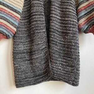May include: A black and grey striped knit cardigan with long sleeves and a ribbed texture. The sleeves have colorful stripes in shades of pink, blue, and beige.