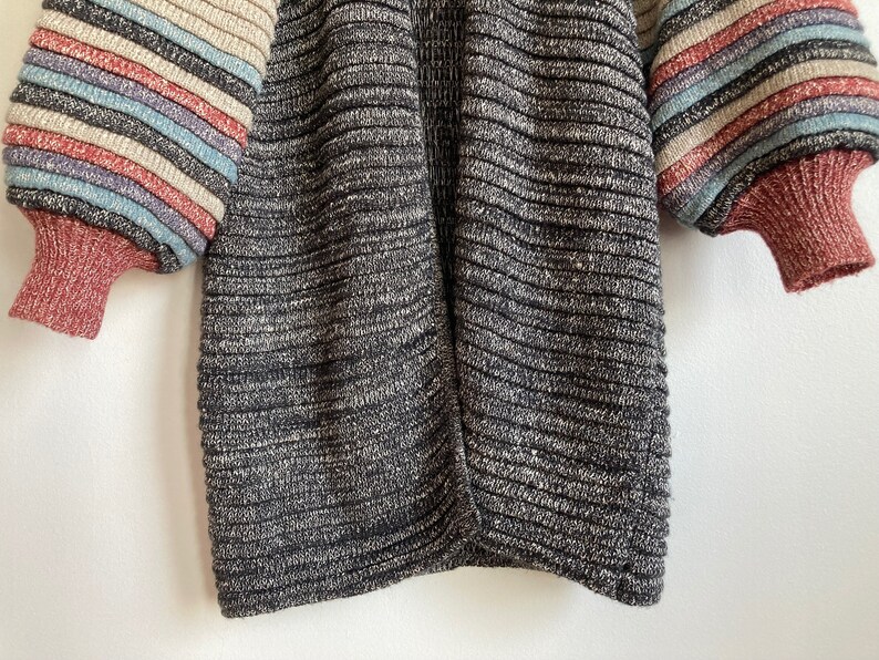 May include: A black and grey striped knit cardigan with long sleeves and a ribbed texture. The sleeves have colorful stripes in shades of pink, blue, and beige.