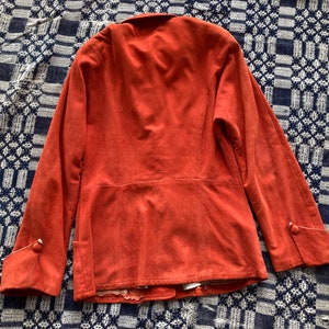 May include: A rust-colored suede jacket with long sleeves and buttoned cuffs. The jacket has a simple design and a slightly fitted silhouette.