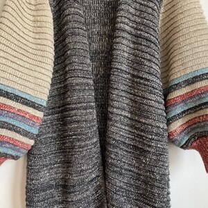 May include: A long-sleeved cardigan with a black and grey knit pattern. The sleeves have a striped pattern in beige, red, blue, and white.