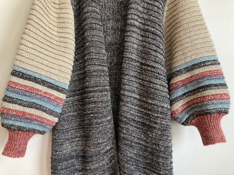 May include: A long-sleeved cardigan with a black and grey knit pattern. The sleeves have a striped pattern in beige, red, blue, and white.