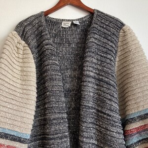 May include: A gray and beige striped cardigan with long sleeves. The sleeves have a beige ribbed texture and a multi-colored stripe at the cuff. The cardigan has a V-neck and a label that reads "home" in the center.