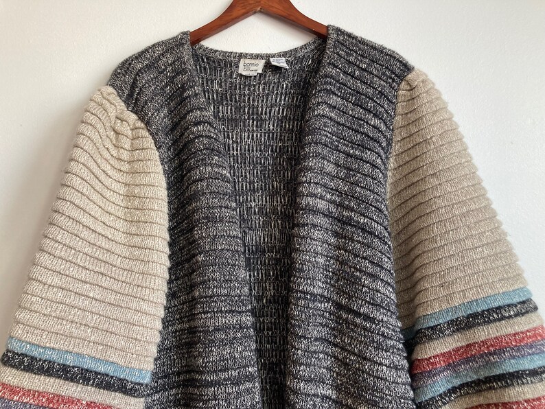 May include: A gray and beige striped cardigan with long sleeves. The sleeves have a beige ribbed texture and a multi-colored stripe at the cuff. The cardigan has a V-neck and a label that reads "home" in the center.