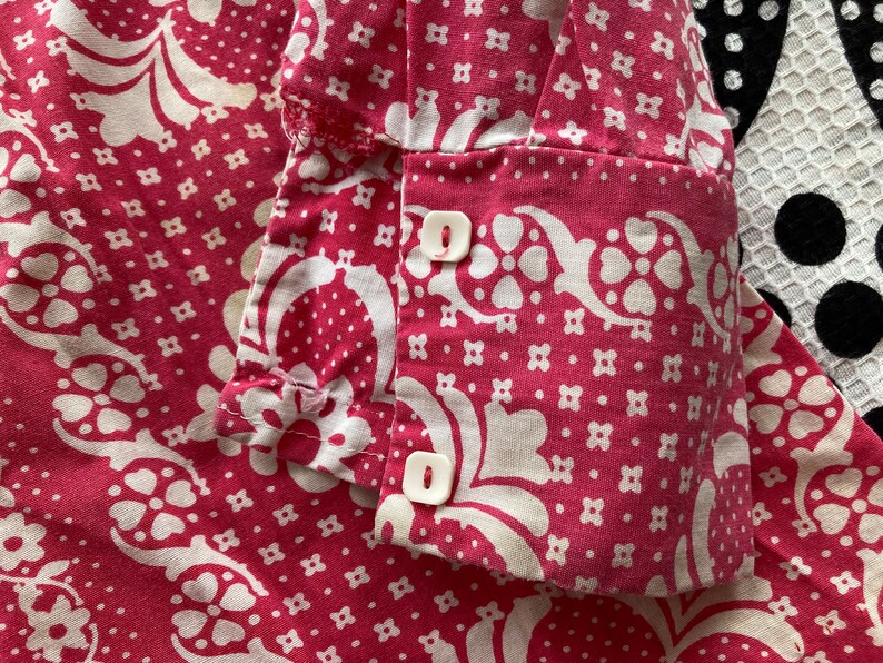 May include: Close-up of a pink fabric with a white floral pattern. The fabric has two white buttons on the cuff.