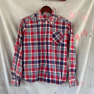 May include: A long-sleeved plaid shirt in red, blue, white, and gray. The shirt has a button-down front, a chest pocket, and buttoned cuffs. The shirt is hanging on a wooden hanger.