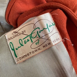 May include: A close-up of a brown suede jacket with a white label that reads "Tailored by Jules-Taylor of Johnstown, New York".
