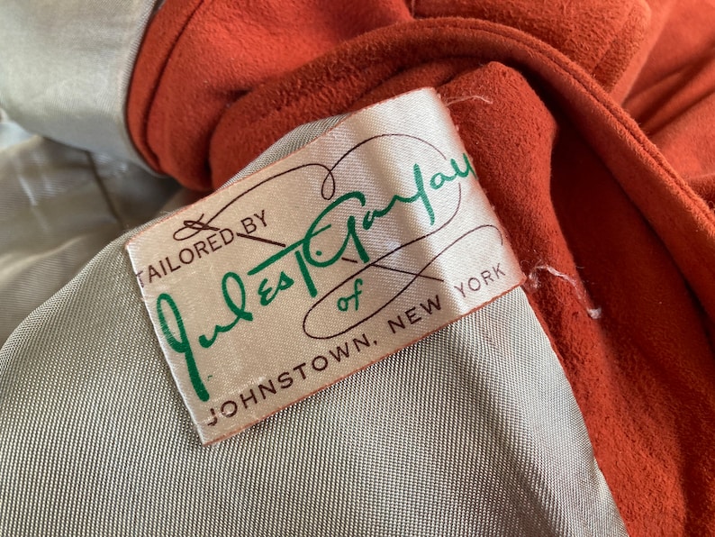May include: A close-up of a brown suede jacket with a white label that reads "Tailored by Jules-Taylor of Johnstown, New York".