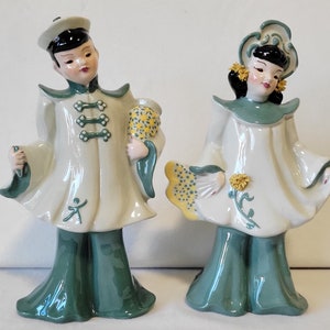 May include: Two ceramic figurines of a man and woman dressed in traditional Chinese clothing. The man is holding a bouquet of flowers and the woman is holding a fan. Both figurines are wearing green and white clothing and have black hair.