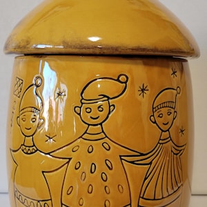 May include: A yellow ceramic cookie jar with a lid shaped like a roof. The jar features three cartoon figures with brown outlines and black eyes. The figures are wearing hats and have smiling faces. The jar has a brown glaze and a glossy finish.