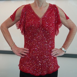 May include: A red sequined top with a V-neck and short, ruffled sleeves. The top has a cut-out detail at the shoulders.