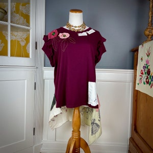 May include: A burgundy t-shirt with a floral applique and a white lace trim. The shirt is on a mannequin and has a high-low hemline.