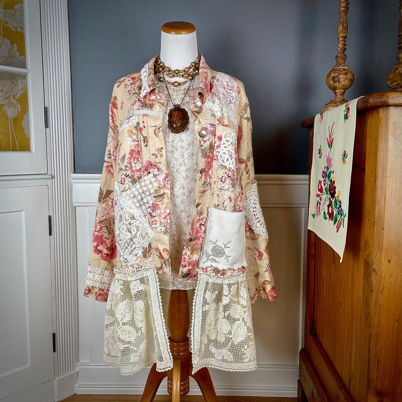 May include: A long, beige floral jacket with pink and red flowers, patchwork lace details, and a lace hem. The jacket is displayed on a mannequin with a necklace and pendant. The jacket has two pockets with embroidered floral designs.