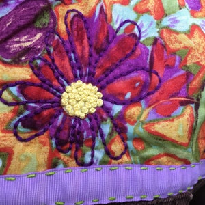 May include: A close-up of a purple and red embroidered flower with a yellow center on a colorful fabric background. The fabric has a purple ribbon sewn to the bottom edge.