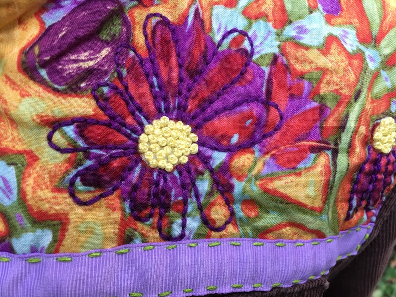 May include: A close-up of a purple and red embroidered flower with a yellow center on a colorful fabric background. The fabric has a purple ribbon sewn to the bottom edge.