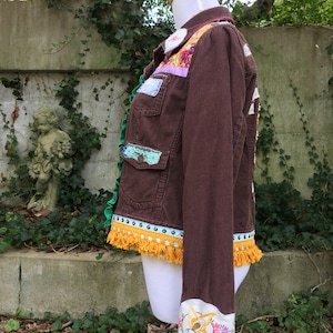 May include: A brown corduroy jacket with colorful patchwork details, including floral embroidery, lace, and fringe. The jacket has a yellow tassel trim and a butterfly embroidered on the cuff.