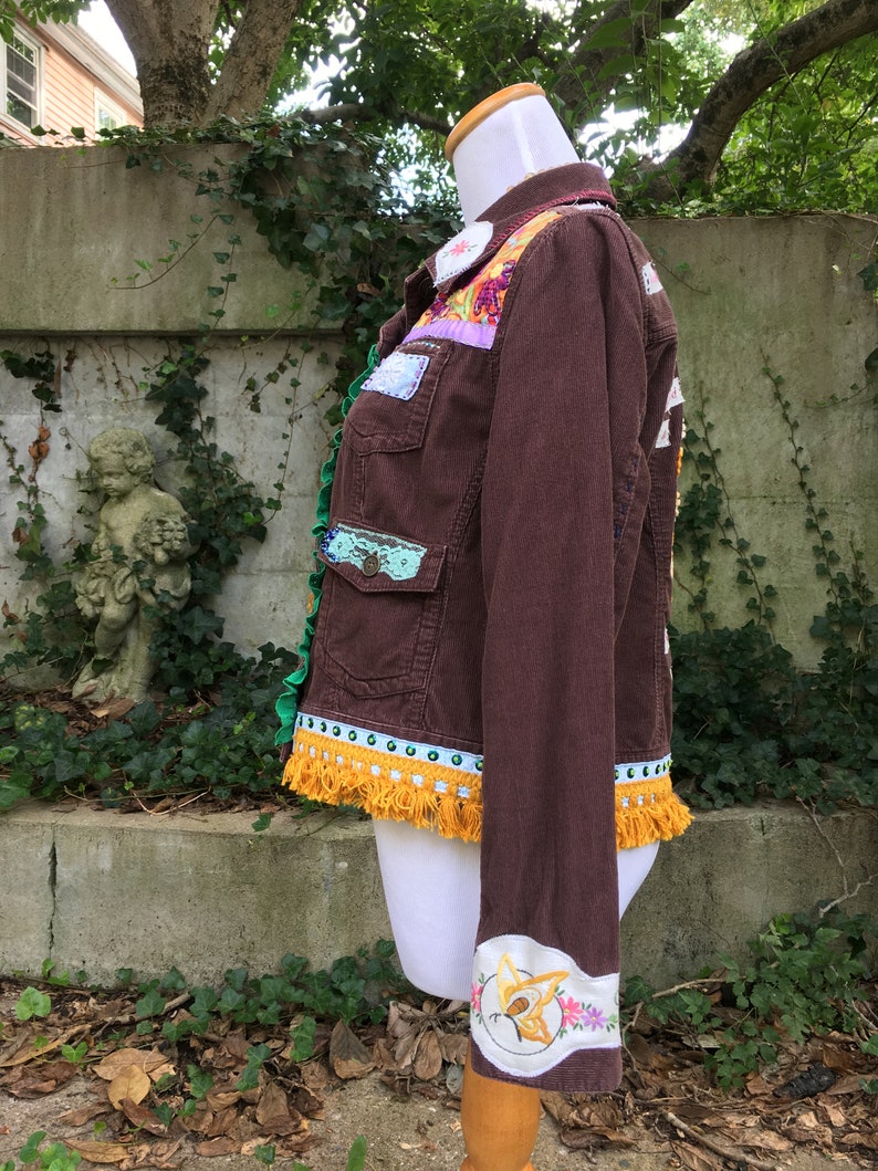May include: A brown corduroy jacket with colorful patchwork details, including floral embroidery, lace, and fringe. The jacket has a yellow tassel trim and a butterfly embroidered on the cuff.