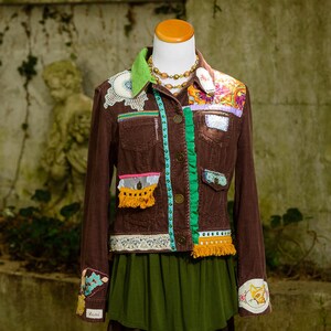 May include: A brown corduroy jacket with colorful fabric embellishments, including lace, ribbon, and fringe. The jacket has a green ribbon down the front and a yellow fringe trim on the pockets.