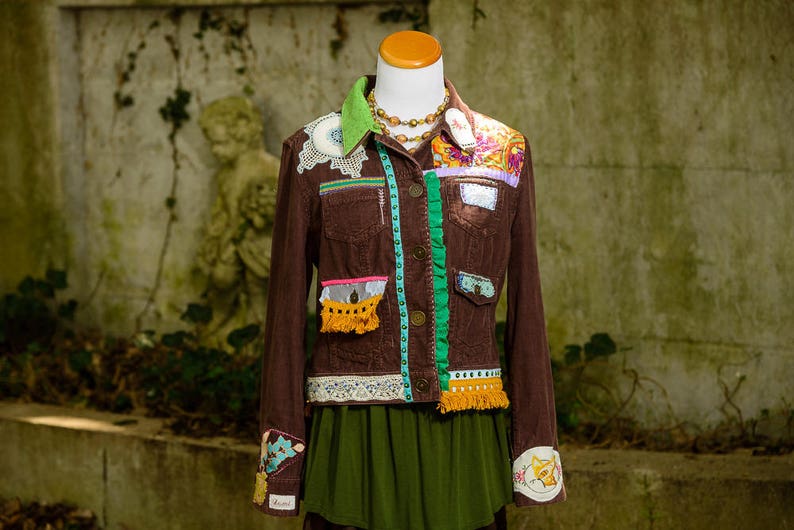 May include: A brown corduroy jacket with colorful fabric embellishments, including lace, ribbon, and fringe. The jacket has a green ribbon down the front and a yellow fringe trim on the pockets.
