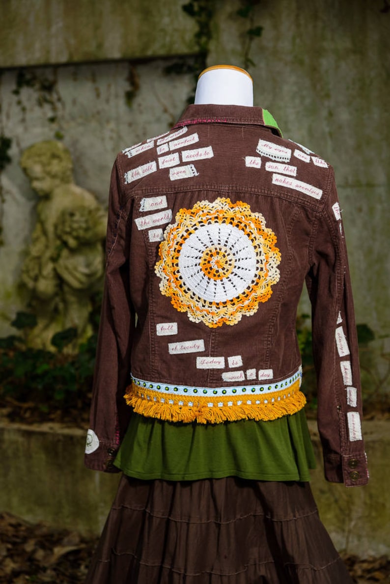 May include: Brown corduroy jacket with a crocheted doily on the back. The jacket is decorated with various fabric scraps with handwritten text, including "the sun that makes armholes" and "full of beauty".