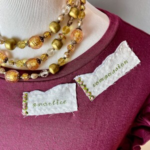 May include: A maroon t-shirt with two white fabric patches sewn on. The patches have green embroidery that reads "practice" and "imagination". The t-shirt has a ruffled trim on the sleeves and a gold beaded necklace is draped over the neckline.