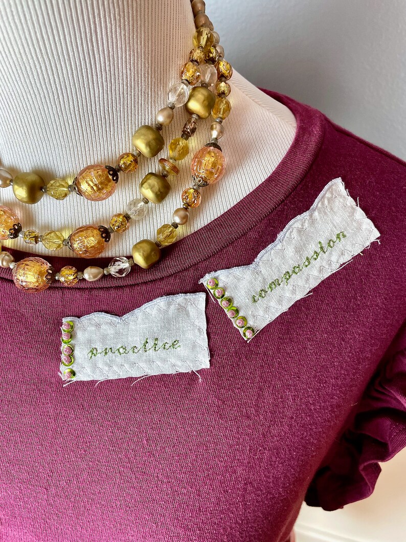 May include: A maroon t-shirt with two white fabric patches sewn on. The patches have green embroidery that reads "practice" and "imagination". The t-shirt has a ruffled trim on the sleeves and a gold beaded necklace is draped over the neckline.
