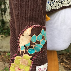 May include: Brown corduroy jacket with floral patches on the sleeves and a white lace trim with blue beads on the collar. The jacket has a small white label with the word "Rustic" on it.