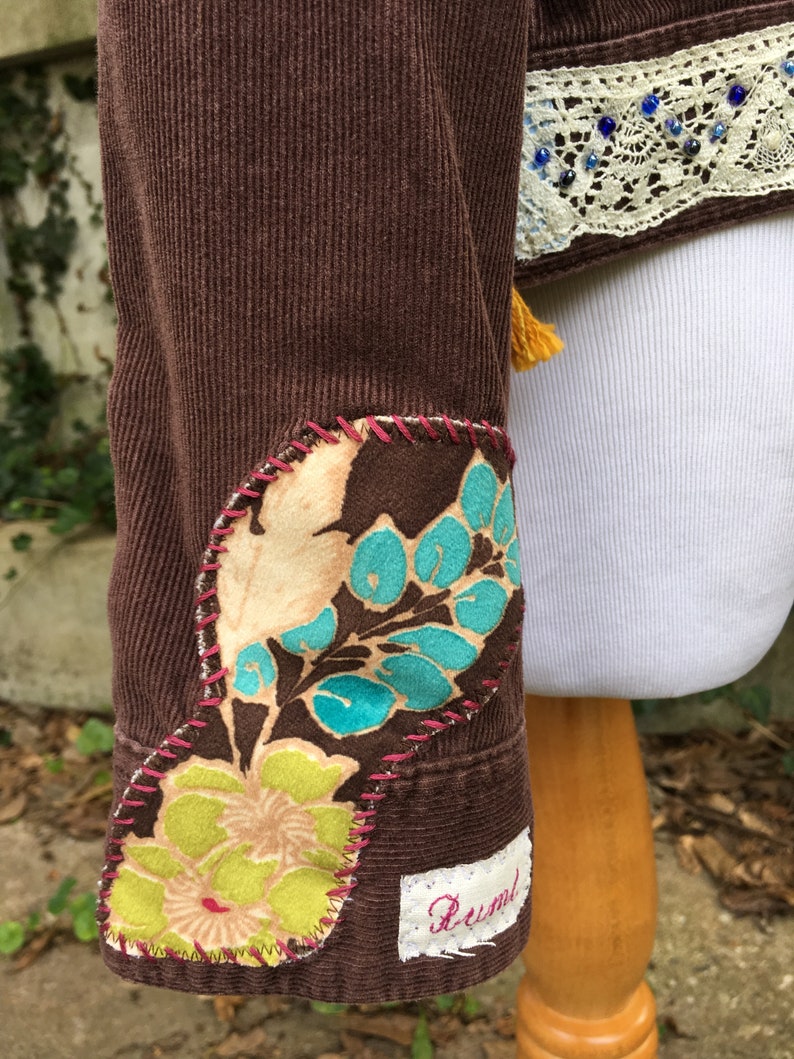 May include: Brown corduroy jacket with floral patches on the sleeves and a white lace trim with blue beads on the collar. The jacket has a small white label with the word "Rustic" on it.
