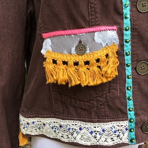 May include: A brown corduroy jacket with a white lace trim, yellow fringe, and turquoise and green beaded trim. The jacket has a pink ribbon and a white lace pocket.