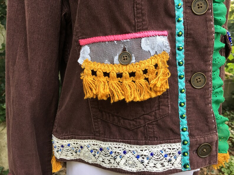 May include: A brown corduroy jacket with a white lace trim, yellow fringe, and turquoise and green beaded trim. The jacket has a pink ribbon and a white lace pocket.