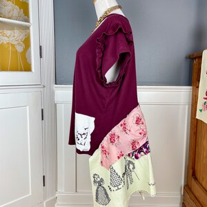 May include: A maroon t-shirt with a ruffled neckline and short sleeves. The shirt is layered with a white lace panel, a pink floral fabric, and a cream fabric with an embroidered design.