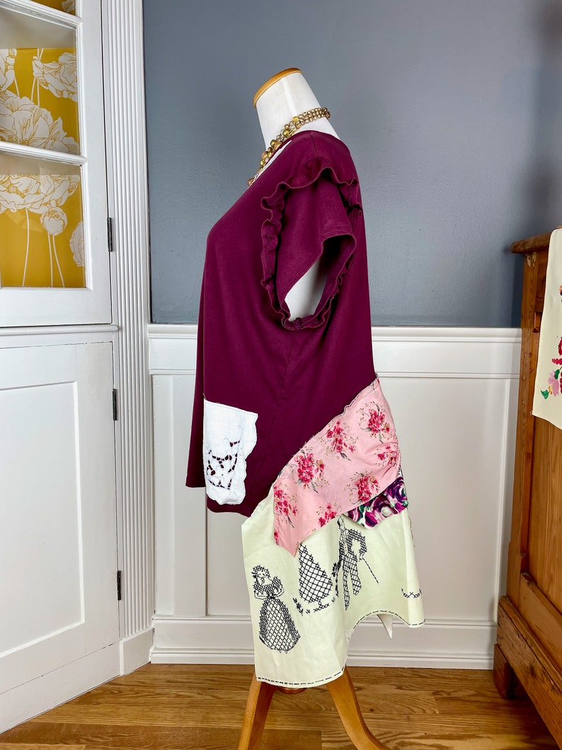 May include: A maroon t-shirt with a ruffled neckline and short sleeves. The shirt is layered with a white lace panel, a pink floral fabric, and a cream fabric with an embroidered design.