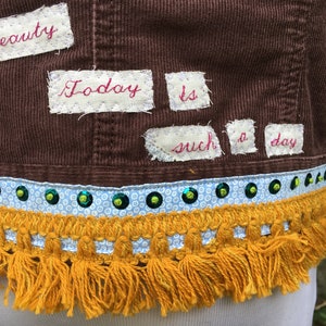 May include: A brown corduroy jacket with a white fabric trim and gold fringe. The jacket has patches sewn on with the words "of beauty", "Today", "is", "such", "a", and "day" written in red thread.