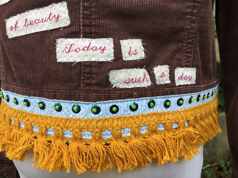 May include: A brown corduroy jacket with a white fabric trim and gold fringe. The jacket has patches sewn on with the words "of beauty", "Today", "is", "such", "a", and "day" written in red thread.