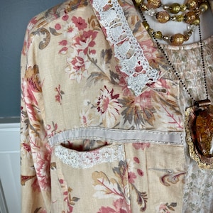 May include: A floral print jacket with a cream-colored base, adorned with pink and burgundy flowers and gray leaves. The jacket features white lace trim on the collar and pocket, and a cream-colored ribbon detail. A layered necklace with gold and amber tones completes the look.