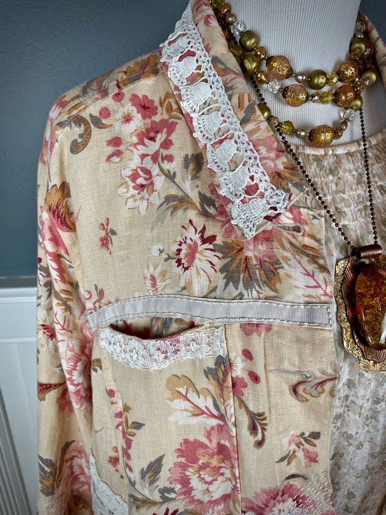 May include: A floral print jacket with a cream-colored base, adorned with pink and burgundy flowers and gray leaves. The jacket features white lace trim on the collar and pocket, and a cream-colored ribbon detail. A layered necklace with gold and amber tones completes the look.