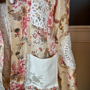 May include: A beige floral jacket with pink and white flower patterns. The jacket features white lace accents on the pockets and sleeves, and decorative buttons. The jacket has a vintage, bohemian style.