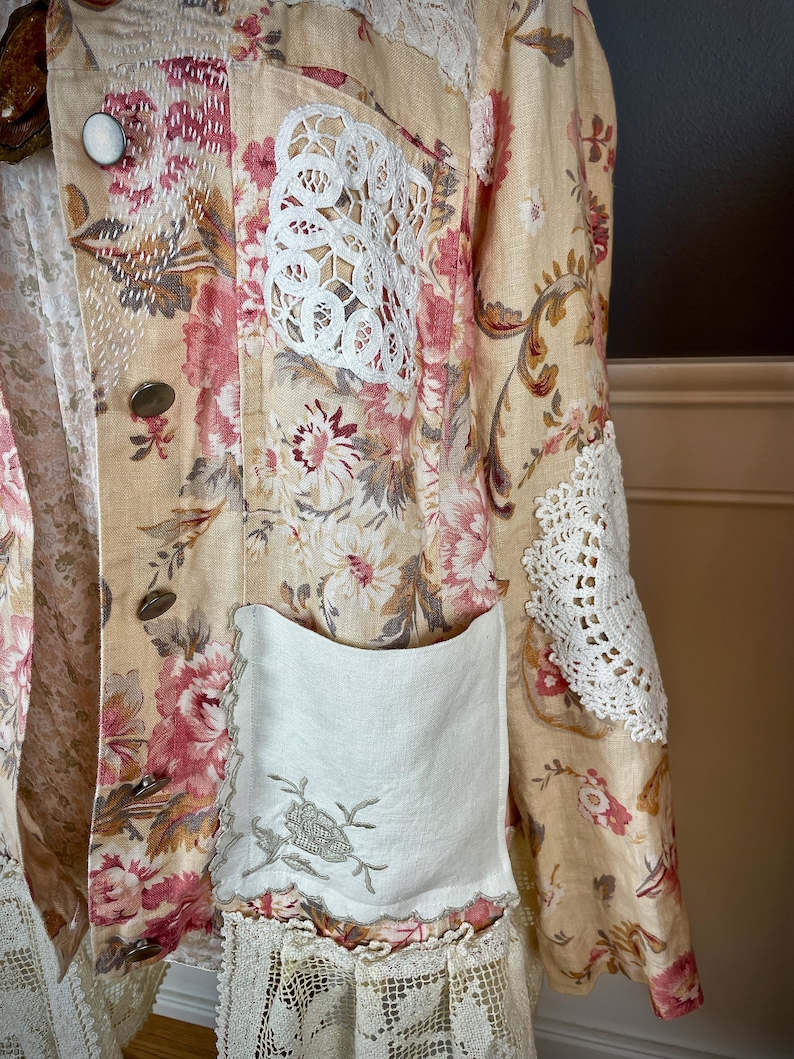 May include: A beige floral jacket with pink and white flower patterns. The jacket features white lace accents on the pockets and sleeves, and decorative buttons. The jacket has a vintage, bohemian style.
