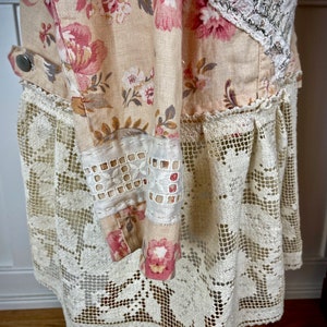 May include: A cream-colored dress with a floral pattern in pink and white. The dress features a lace overlay at the hem and sleeves, with a button detail on the side. The fabric has a vintage, handmade aesthetic.