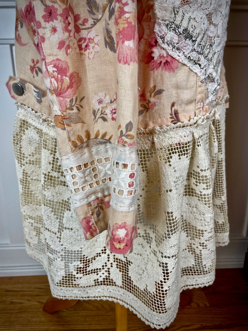 May include: A cream-colored dress with a floral pattern in pink and white. The dress features a lace overlay at the hem and sleeves, with a button detail on the side. The fabric has a vintage, handmade aesthetic.