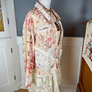 May include: A floral print jacket with lace trim and a matching lace skirt. The jacket features a cream-colored base with pink and red floral patterns. The jacket and skirt are displayed on a mannequin with a wooden stand.