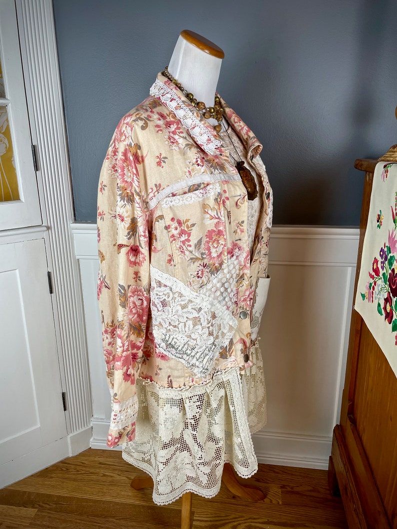May include: A floral print jacket with lace trim and a matching lace skirt. The jacket features a cream-colored base with pink and red floral patterns. The jacket and skirt are displayed on a mannequin with a wooden stand.