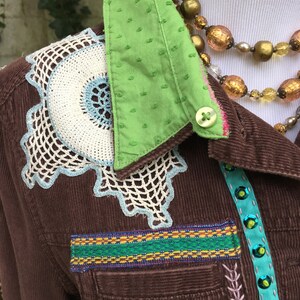 May include: A brown corduroy jacket with a green polka dot collar, white and blue crocheted embellishments, and a colorful ribbon trim. The jacket has a button closure and a pocket on the left side.