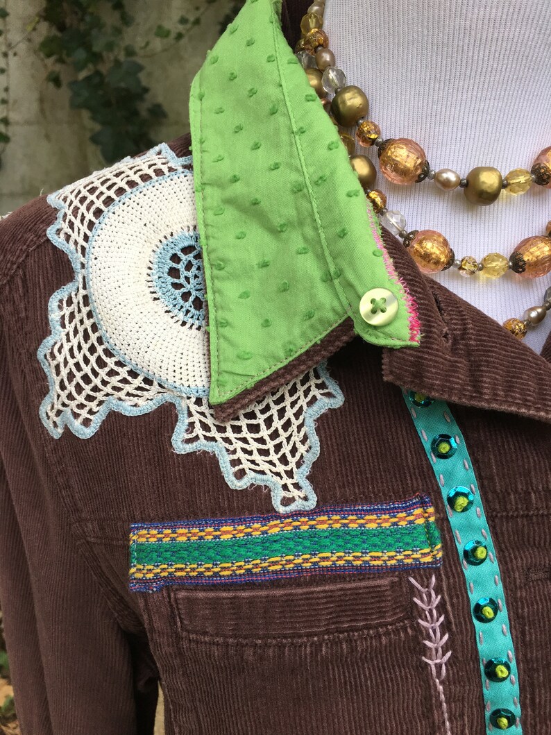 May include: A brown corduroy jacket with a green polka dot collar, white and blue crocheted embellishments, and a colorful ribbon trim. The jacket has a button closure and a pocket on the left side.