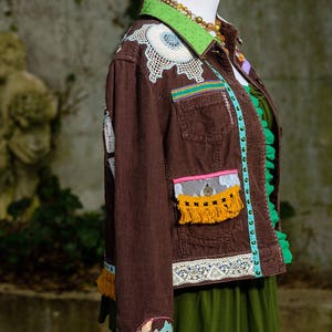 May include: A brown corduroy jacket with a green collar and lace trim. The jacket is decorated with colorful embroidery, beads, and fringe.