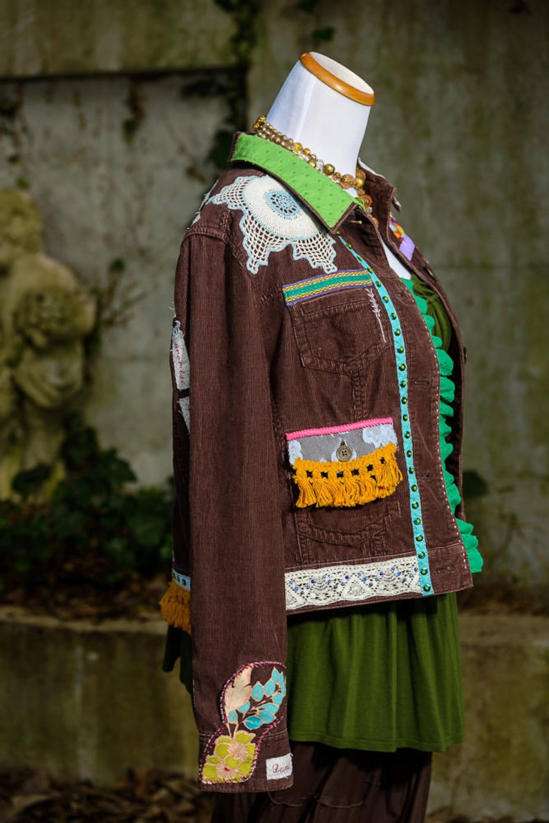 May include: A brown corduroy jacket with a green collar and lace trim. The jacket is decorated with colorful embroidery, beads, and fringe.