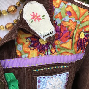 May include: A brown corduroy jacket with a floral fabric patch on the shoulder, a white fabric patch with a pink flower embroidered on it, and a purple ribbon trim. The jacket has a green lace trim on the bottom left side and a white lace patch with a white flower on the bottom right side.