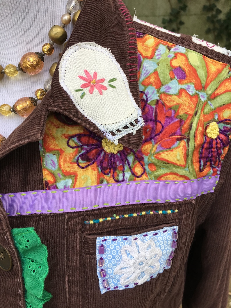 May include: A brown corduroy jacket with a floral fabric patch on the shoulder, a white fabric patch with a pink flower embroidered on it, and a purple ribbon trim. The jacket has a green lace trim on the bottom left side and a white lace patch with a white flower on the bottom right side.