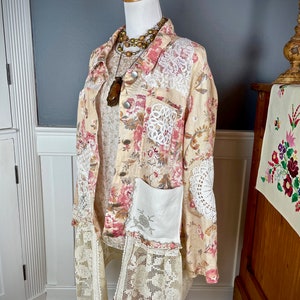 May include: A beige floral print jacket with pink and red flowers, white lace accents, and a white pocket. The jacket is displayed on a mannequin with a layered necklace and a wooden pendant. The jacket has a lace trim at the bottom.